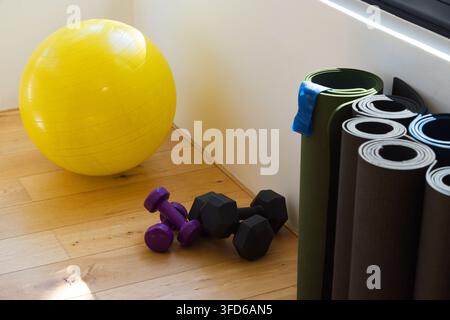 Balle de stabilité jaune reposant sur un sol en bois à gauche, haltères violets allongés, tapis empilés, espace de copie Banque D'Images