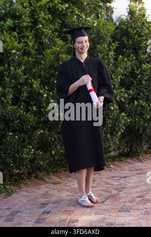 Diplômée portant une toque et une robe tenant un diplôme avec un ruban rouge sur une cour en briques, souriante Banque D'Images