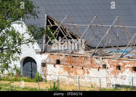 Bâtiment en briques abandonné avec un toit effondré, des poutres en bois apparentes, des murs cassés, une clôture métallique et des arbres verts par une journée ensoleillée. Banque D'Images