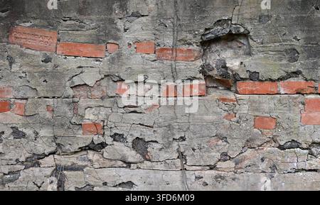 Surface murale en ciment décrépie avec enduit fissuré et altéré, brique rouge cassée exposée, sous-couche sombre Banque D'Images