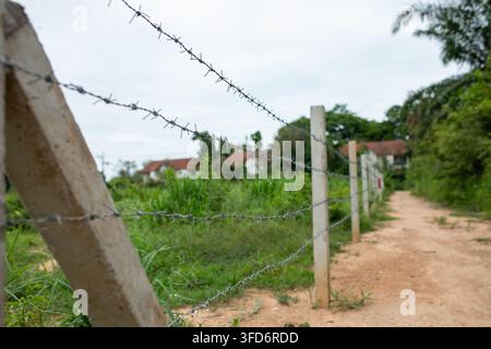 Clôture en fil de fer barbelé avec poteaux en béton bordant un chemin de terre à côté d'un champ envahi par la végétation, maisons rurales avec toits rouges visibles à travers les arbres verts sous un ciel nuageux Banque D'Images