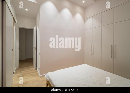 Chambre moderne minimaliste avec lit blanc, parquet clair, placards encastrés et éclairage encastré doux dans un intérieur résidentiel contemporain calme Banque D'Images