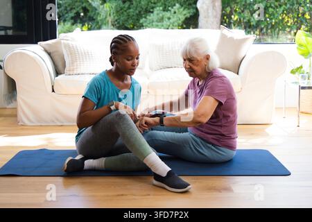 Femme afro-américaine et femme âgée en tenue de sport vérifiant des appareils de poignet sur un tapis bleu à la maison Banque D'Images