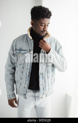 Un jeune homme élégant à la peau foncée et aux cheveux bouclés pose dans un décor de studio, sa main près de son cou Banque D'Images