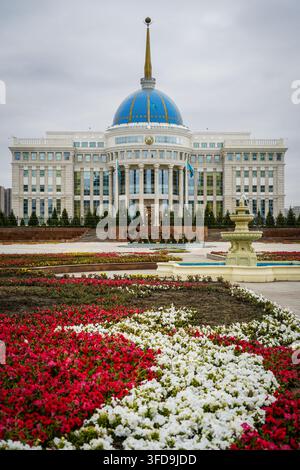 Le palais présidentiel Ak Orda, un grand bâtiment blanc avec un dôme bleu proéminent et une flèche dorée, se dresse sous un ciel nuageux. Au premier plan, rouge Banque D'Images