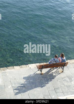CHANIA, CRÈTE - 5 MAI 2025 - Couple romantique se relaxant sur un banc en bois, contemplant les eaux turquoise de l'océan sous un soleil éclatant Banque D'Images