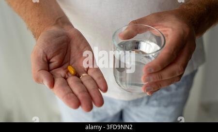 Close-up d'un homme tenant une capsule jaune dans sa main et un verre d'eau, prêt à prendre un médicament ou un complément alimentaire. Banque D'Images