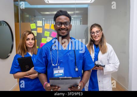 Équipe médicale diversifiée dans une clinique moderne, médecin souriant avec tableau de bord se tient entre deux collègues féminines en blouse et manteau de laboratoire, confiant et collégial Banque D'Images