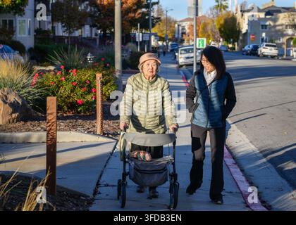 Sénior accompagnée de sa fille adulte, marchant avec une marchette de mobilité. Le soleil d'automne projette de longues ombres. Californie. Banque D'Images