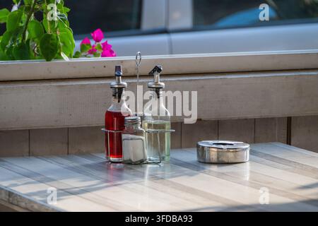 Un set de cruette de table contenant des bouteilles d'huile et de vinaigre rouge, un salière et un cendrier en métal sont posés sur une table de restaurant extérieure sous une lumière douce. Banque D'Images