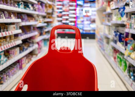 Poignée de chariot rouge avançant dans les allées d'un magasin remplies de produits sur des étagères lumineuses Banque D'Images