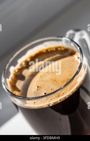 Prise en gros plan extrême de la couche de mousse dorée de la crème sur un espresso noir fraîchement préparé dans une tasse transparente baignée de lumière naturelle. Banque D'Images