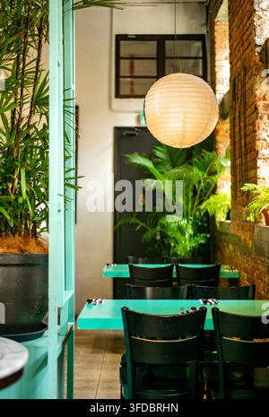 Une photographie d'intérieur atmosphérique d'une charmante salle à manger de restaurant présentant des tables turquoise disposées avec des couverts, des chaises en bois sombre, des plantes en pot des tropiques Banque D'Images