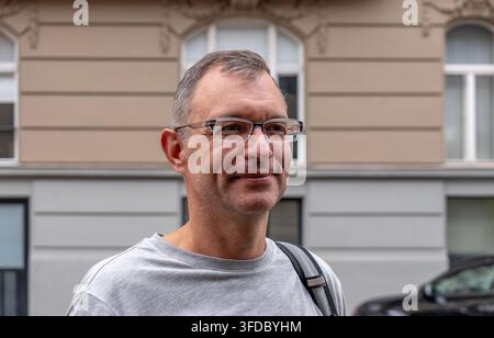 Un homme souriant d'âge moyen portant des lunettes se tient à l'extérieur devant une façade de bâtiment. Banque D'Images