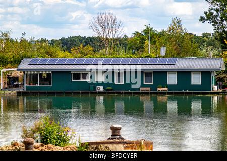 Maison flottante aux murs verts, panneaux solaires sur le toit et petites fenêtres, petite terrasse, arbres aux feuilles d'automne en arrière-plan, journée ensoleillée à Maastr Banque D'Images