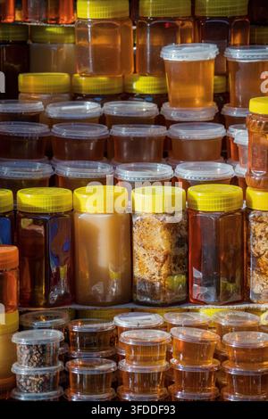 De nombreux bocaux en plastique transparent et en verre remplis de différents types de miel, certains contenant des rayons de miel avec de la propolis, sont empilés en rangées sur un étal de marché Banque D'Images