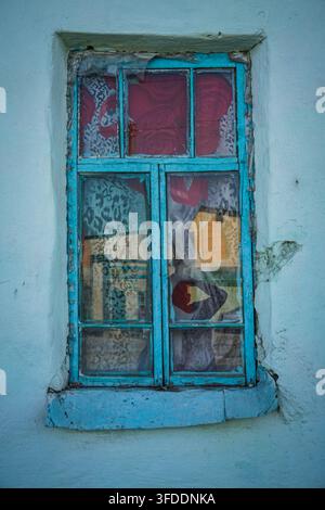 Un plan rapproché d'une vieille fenêtre à quatre panneaux avec un cadre et un appui en bois peint en bleu-vert écaillé, posée contre un mur en plâtre bleu clair rugueux. Beh Banque D'Images