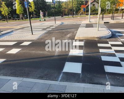 Un passage piéton nouvellement peint avec des rayures blanches audacieuses sur l'asphalte sombre, comportant une îlot de trafic avec revêtement tactile. En arrière-plan, un Banque D'Images