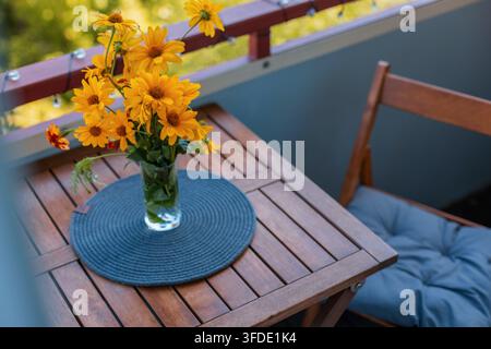 Bouquet de fleurs jaune vif dans un vase en verre sur une table en bois de balcon, représentant un cadre de patio extérieur confortable avec une lumière chaude et une banquette en bois Banque D'Images