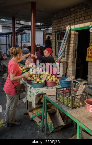 Bichkek, Kirghizistan - 17 septembre 2017 : Un vendeur portant un foulard rouge et un tablier remet un sac de pommes à une cliente en chemise rouge sur un marché Banque D'Images