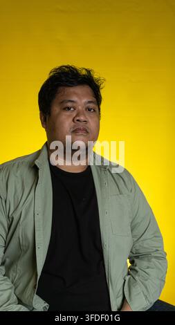 Un portrait décontracté met en scène un jeune homme asiatique avec une expression sérieuse, debout devant un fond jaune vif et uni. Le sujet porte un Banque D'Images
