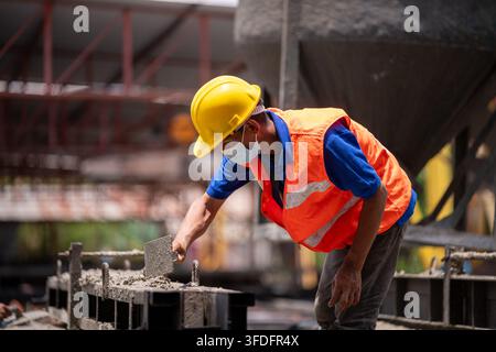 Ouvrier du bâtiment portant un casque de sécurité, un gilet de sécurité et un masque facial lissant le béton humide sur le chantier Banque D'Images