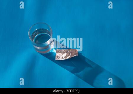 Disposition horizontale plate d'un verre d'eau propre et d'un blister de médicaments vide et utilisé sur une surface bleue. Banque D'Images