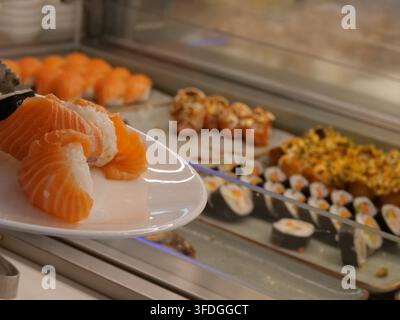 Prise de vue concentrée sur un délicieux sushi nigiri au saumon servi dans une assiette en porcelaine blanche tenue par un client. Le fond flou montre une grande variété de fré Banque D'Images