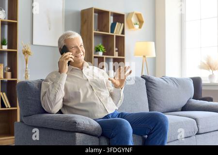 Homme senior heureux parlant au smartphone à la maison, faisant un signe de la main, assis sur le canapé Banque D'Images