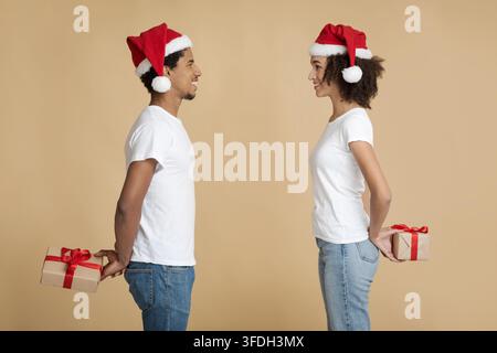 Jeune homme et femme souriant dans le père Noël chapeau look les uns aux autres et de tenir des cadeaux surprise derrière le dos Banque D'Images