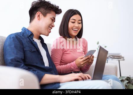 Couple asiatique utilisant un ordinateur portable et un smartphone assis sur un canapé à l'intérieur Banque D'Images