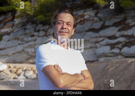 Un homme d'âge moyen aux bras croisés se tient à l'extérieur près d'un mur de pierre baigné de soleil en Croatie. Banque D'Images