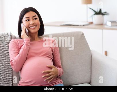 Bonne femme enceinte asiatique parlant au téléphone assis à la maison Banque D'Images