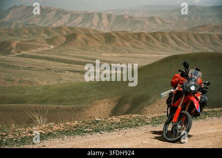 Vue d'une moto d'aventure rouge garée sur un sentier de terre surplombant les vastes collines ondulantes et le paysage montagneux de Song-Kul, région de Naryn, Kirghizistan Banque D'Images