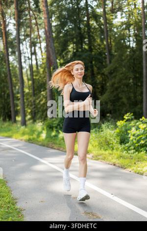 Prise en vue en pleine longueur d'une femme athlétique rousse courant le long d'un chemin de parc, ses longs cheveux volant au vent. Vêtue de vêtements de sport noirs, profitant d'une voiture ensoleillée Banque D'Images