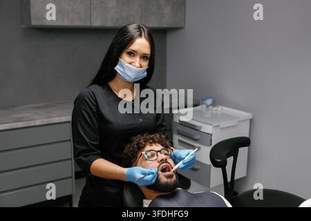 Dentiste femme portant un masque et des gants effectuant un bilan dentaire sur un patient masculin dans une clinique moderne. Se concentrer sur l'hygiène bucco-dentaire Banque D'Images