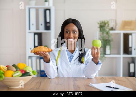 Faire des choix alimentaires sains. Belle nutritionniste féminine tenant croissant et pomme à table dans le bureau médical Banque D'Images