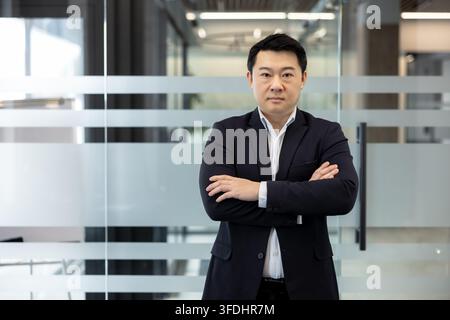 Homme d'affaires asiatique debout avec assurance, les bras croisés, regardant la caméra dans un bureau d'entreprise moderne, représentant le leadership, le succès et le professionnalisme dans un environnement commercial Banque D'Images