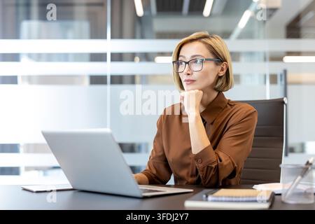 Femme d'affaires portant des lunettes assise à un bureau avec un ordinateur portable, regardant au loin et réfléchissant à la stratégie, au leadership et au développement professionnel dans un environnement d'entreprise Banque D'Images