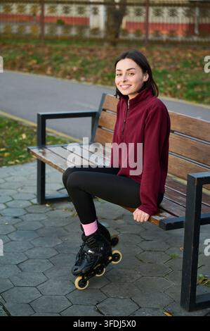 Femme assise sur un banc en bois en patins à roulettes, souriant à l'objectif en polaire bordeaux et leggings noirs. Convient pour les thèmes sportifs, de branding de mode de vie, de confiance ou d'activités du week-end Banque D'Images