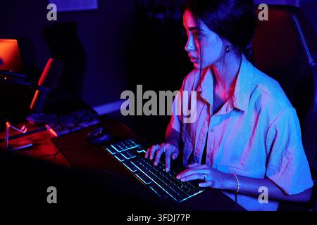 Femme utilisant une configuration de jeu avec un clavier rétroéclairé dans une pièce sombre, illuminée par une lumière rouge et bleue néon Banque D'Images