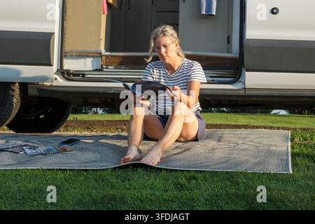 Une femme senior détendue est assise pieds nus sur un tapis d'extérieur en train de lire un magazine à côté de son camping-car, entourée d'herbe verte et de la lumière chaude de l'heure dorée Banque D'Images