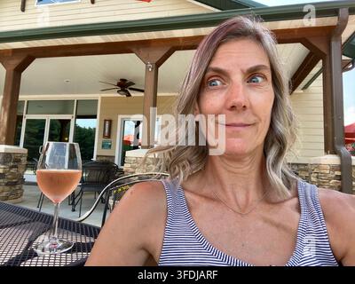 Une femme arborant une expression espiègle et surprise est assise à une table extérieure sur la terrasse d'un domaine viticole, un verre de vin rosé rose devant elle et la dégustation Banque D'Images
