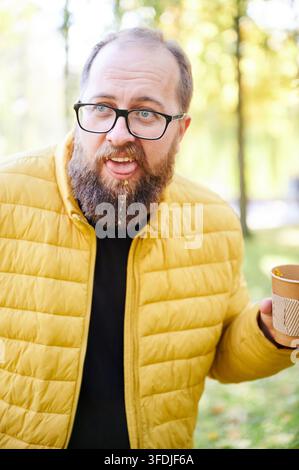 Homme décontracté en veste jaune avec gobelet en papier. Scène urbaine mettant en scène un homme avec un café dans un cadre de parc. Une personne portant une veste jaune tenant négligemment un gobelet en papier dans une atmosphère de parc urbain Banque D'Images