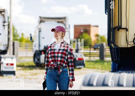 Une conductrice de camion portant une chemise à carreaux rouges et une casquette de baseball regarde au loin tout en se tenant entre deux gros semi-remorques Banque D'Images