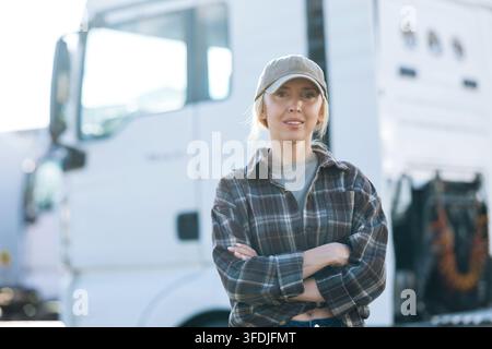 Une conductrice de camion confiante se tient les bras croisés, vêtue d'une chemise à carreaux et d'une casquette, avec un gros semi-remorque derrière elle Banque D'Images