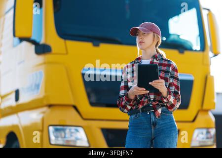 Une femme portant une casquette de baseball et une chemise à carreaux tient une tablette tout en se tenant à côté d'un grand semi-remorque jaune Banque D'Images