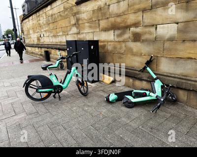 Liverpool, Royaume-Uni - 16 mai 2026 : Scooter électrique renversé et vélo de location sans station appuyés contre un mur en grès sur un trottoir de Liverpool, Royaume-Uni. Banque D'Images