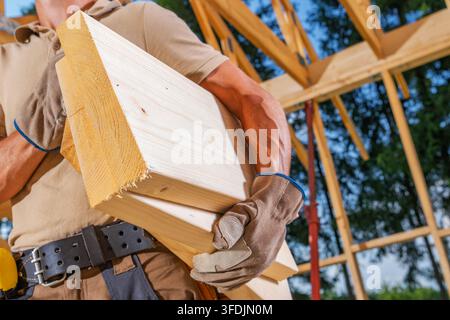 Vue rapprochée d'un charpentier portant une poutre en bois coupée sous un bras. Des gants de travail et une ceinture à outils sont visibles contre la charpente du toit apparente. Banque D'Images