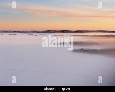 Vue aérienne d'un vaste paysage enneigé avec une brume dense s'élevant au-dessus d'un terrain gelé et de collines lointaines sous un ciel doux au coucher du soleil à Inari, Laponie, F Banque D'Images